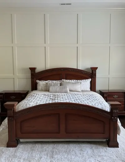A bedroom with a large wooden bed, white bedding, matching wooden nightstands, a dresser, and a white area rug on dark hardwood floors. Light enters through a window on the right.